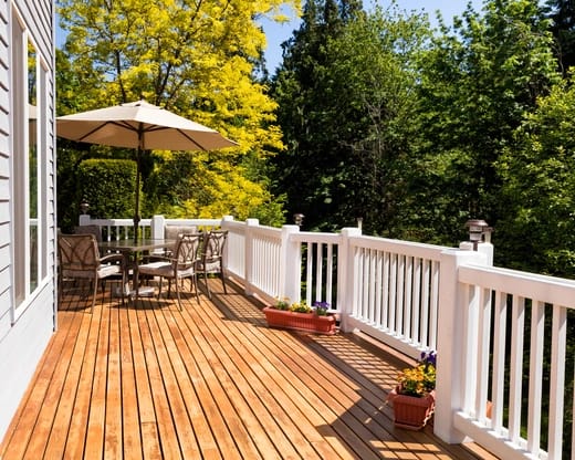 An expansive hardwood deck featuring white balustrade railings, a round patio dining table with chairs, and an umbrella, surrounded by lush trees.