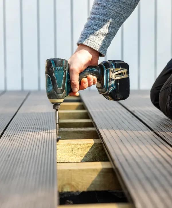 A person drills into wood while installing a deck, focused on his task in a sunny outdoor setting. A person drills into wood while installing a deck, focused on his task in a sunny outdoor setting.